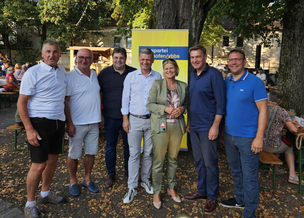 Sechs Männer und eine Frau sehen vor einem Wahlplakat in einem Gastgarten